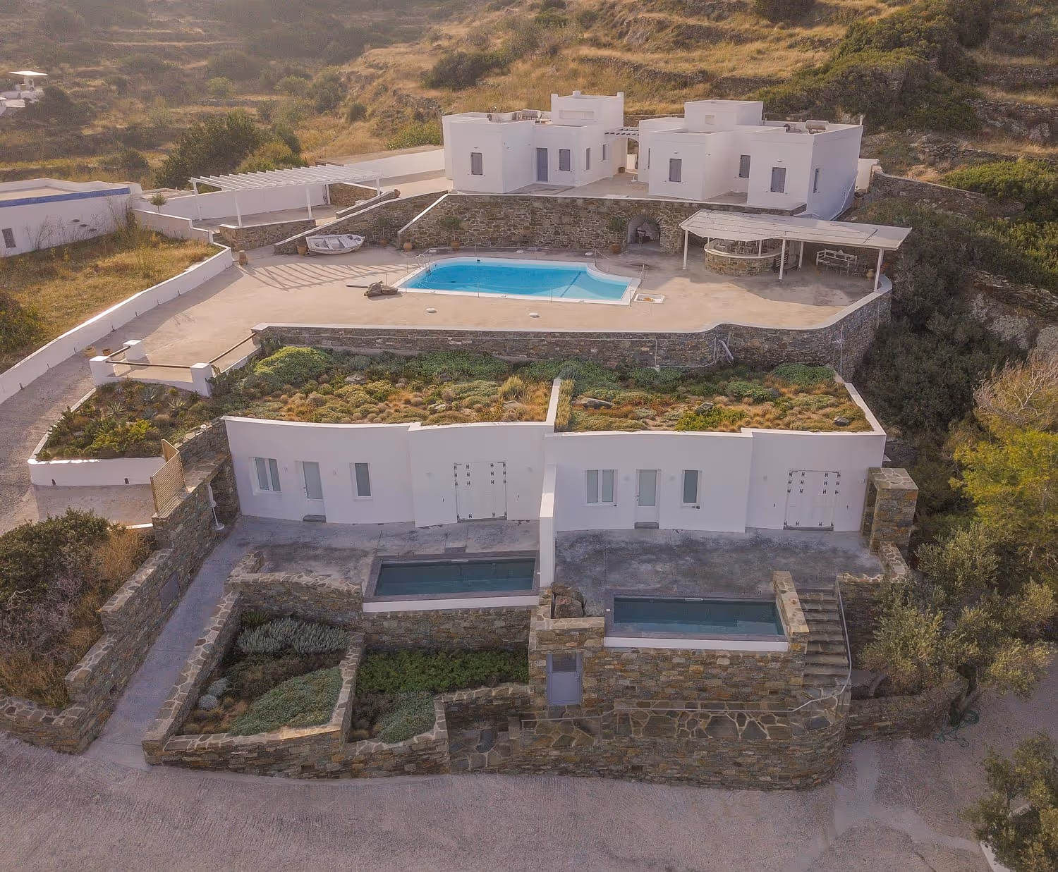 Aerial view of a multi-level white villa with rooftop gardens, stone walls, two small rectangular pools, and a larger swimming pool on the upper terrace.